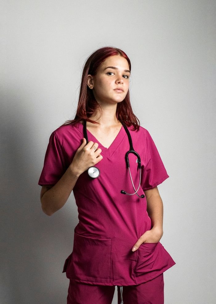 Portrait of a young female doctor in pink scrubs holding a stethoscope, set against a gray background.