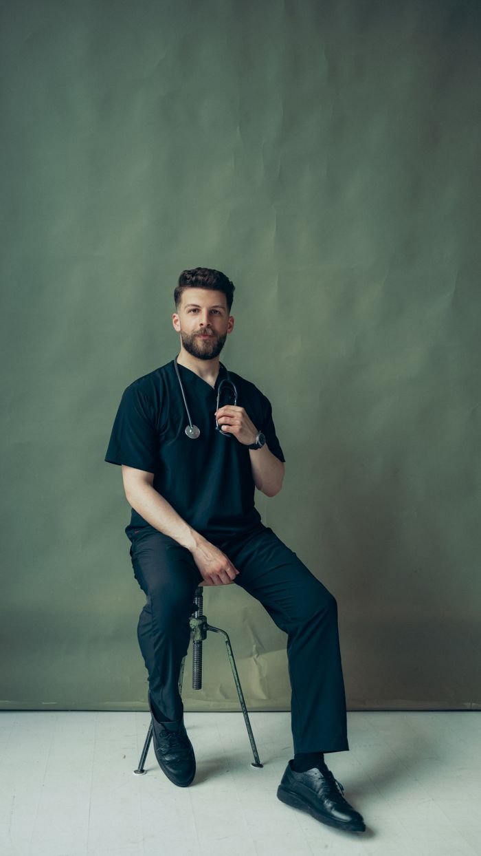 Portrait of a young male doctor sitting indoors with a stethoscope, wearing a healthcare uniform.