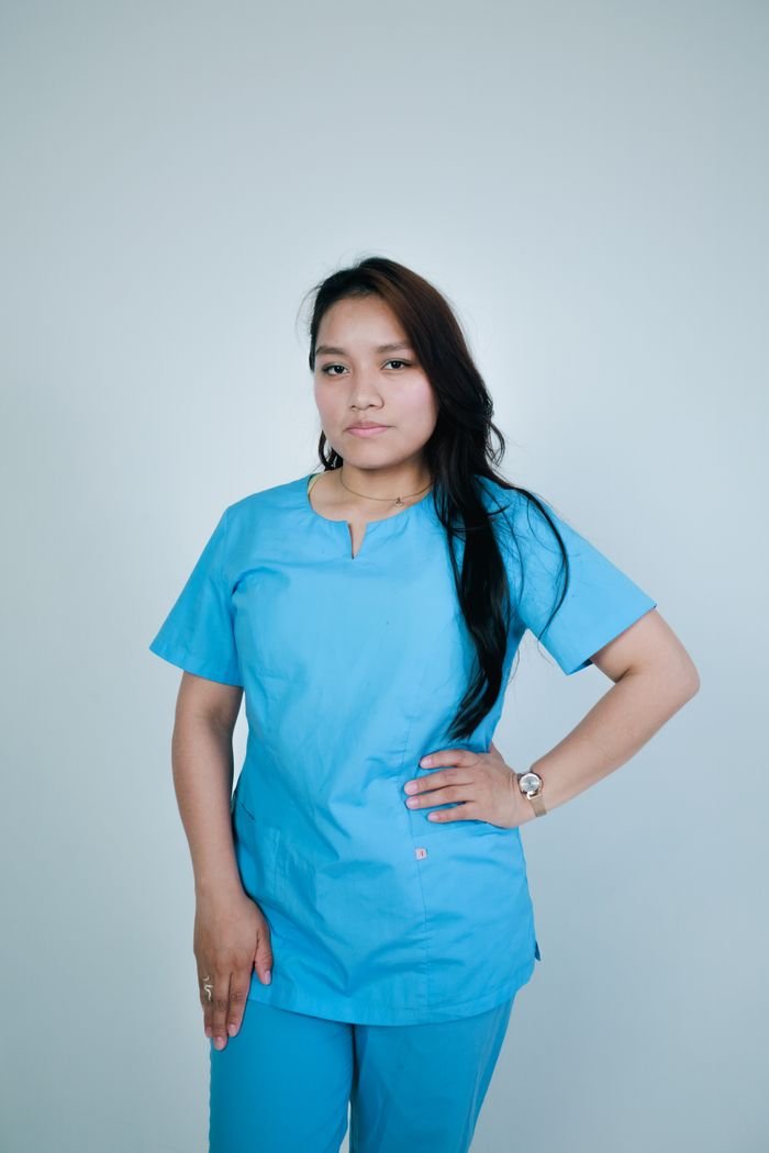 A confident nurse in blue scrubs stands indoors against a neutral background, embodying professionalism.