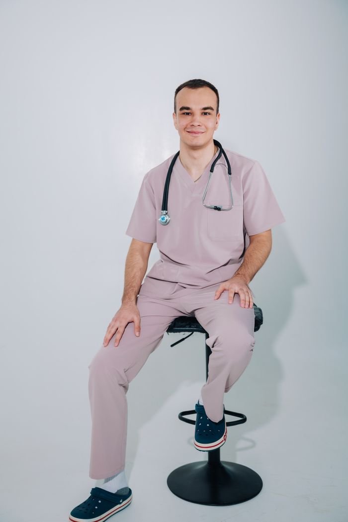 Medical professional sitting on stool in scrubs with stethoscope, isolated on white background.